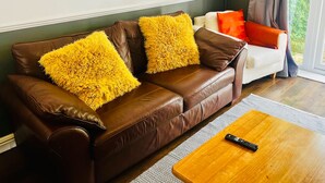 A brown leather sofa with two yellow fluffy pillows, a remote control on a wooden coffee table, and a framed cityscape painting on the wall. - Impeccable 3-bed House in Nottingham Free Parking (Nottingham)