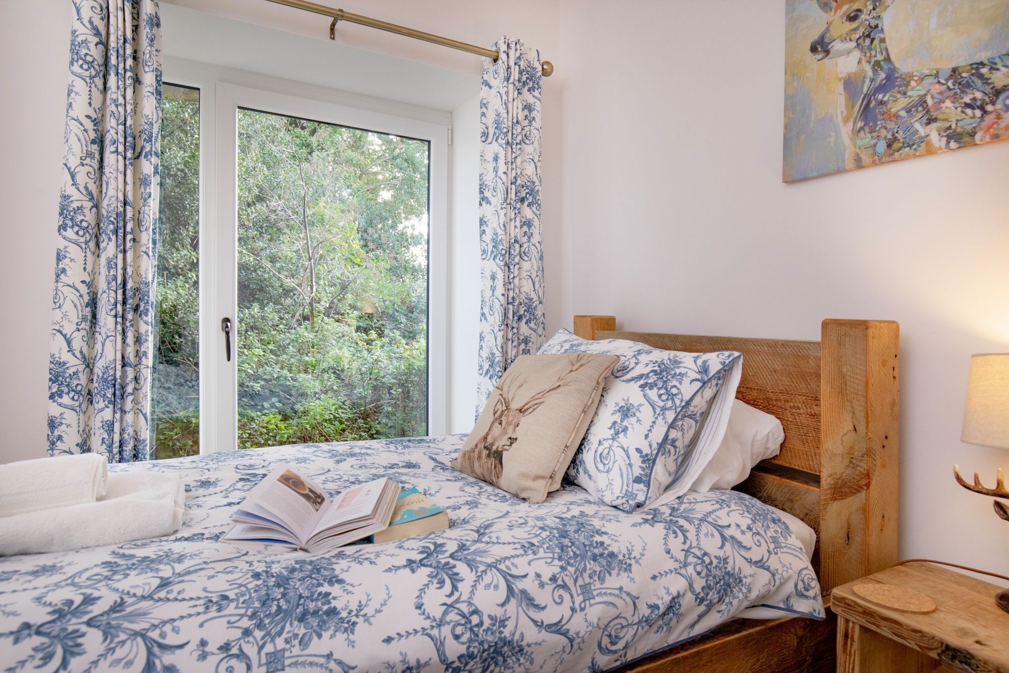 Une chambre avec une grande fenêtre, un lit recouvert d’une couvre-lit à motifs floraux, une tête de lit en bois, une table de chevet avec une lampe et un tableau accroché au mur.