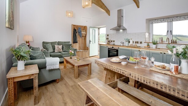A cozy living room with a green sofa, wooden coffee table, and a dining area with a wooden bench and table. - Converted Barn with amazing views - sleeps 4 (Paignton)