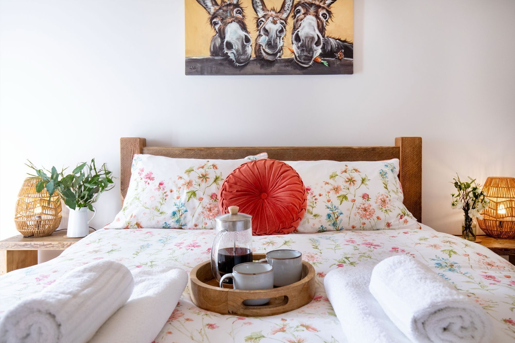 Ein ordentlich bezogenes Bett mit Blumenmuster, ein rotes Zierkissen und ein Tablett mit Teekanne und Tassen.