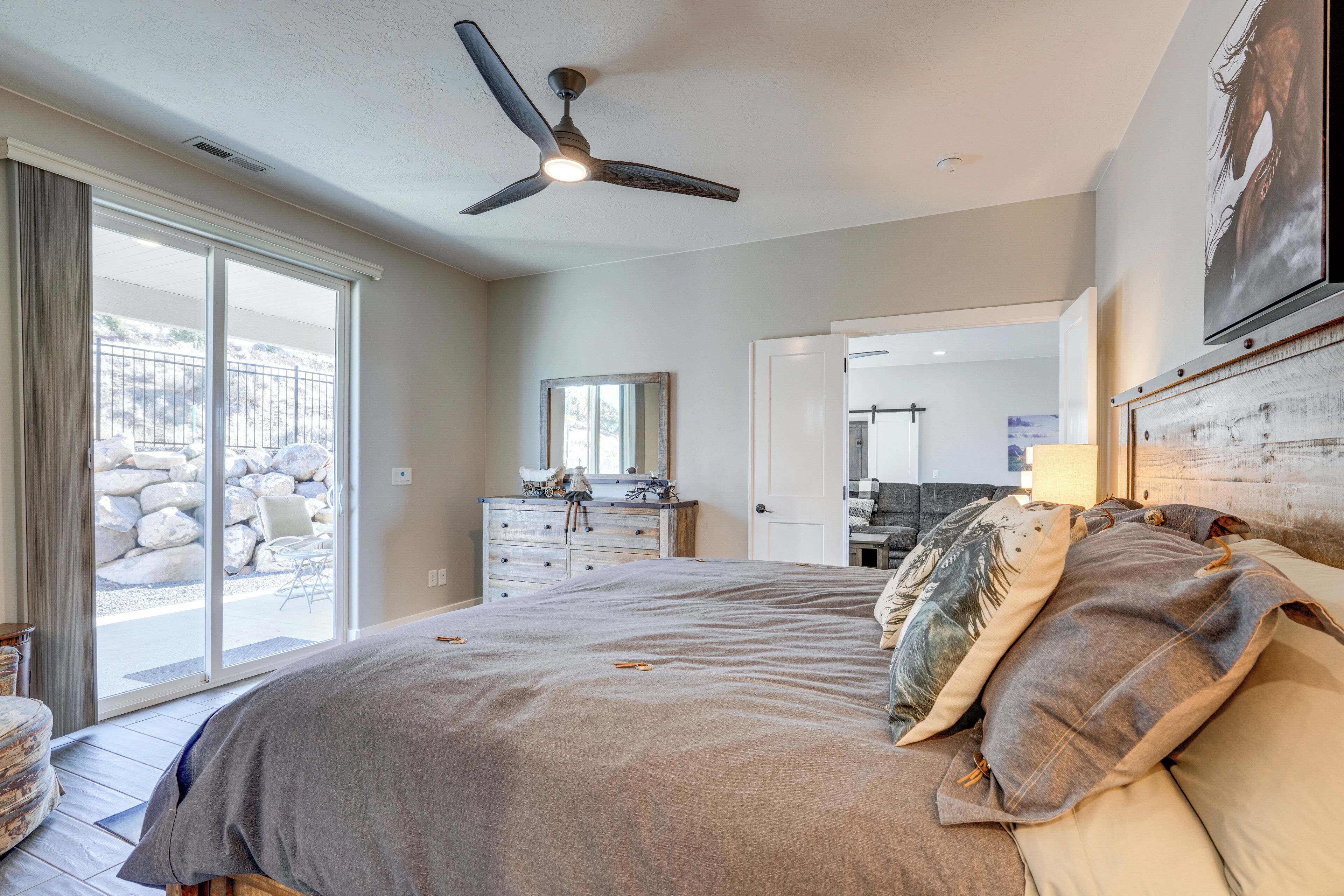 A bedroom with a large bed, a ceiling fan, a dresser, and a view of a stone patio.