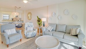 A living room with a grey sofa, two armchairs, a coffee table, a ceiling fan, a dining area with a table and chairs, and a kitchen in the background. - Orange Beach Condo w/ Beach+ Tennis Court Access (Orange Beach)
