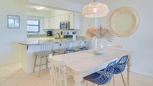 A modern kitchen with white cabinets, a stainless steel refrigerator, and a microwave. There is a dining area with a white table and blue chairs. - Orange Beach Condo w/ Beach+ Tennis Court Access (Orange Beach)