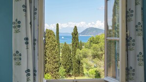 A view through an open window with floral curtains, overlooking a coastal landscape with cypress trees and a distant mountain. - Casa del Duca 10, Emma Villas (Portoferraio)