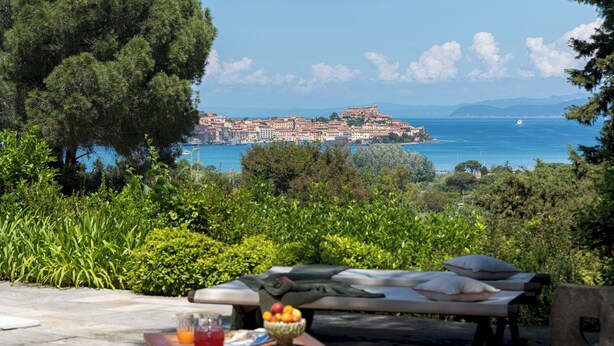 A seaside view with a dining area, outdoor seating, and a distant view of a coastal town. - Casa del Duca 10, Emma Villas (Portoferraio)