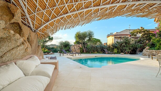 A pool area with a thatched roof, outdoor seating, and a view of surrounding buildings.