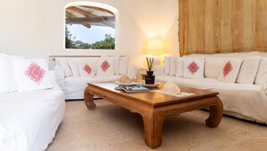 A cozy room with a wooden coffee table, white sofas with decorative pillows, a lamp, and a view through a window.