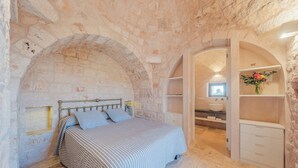 Een slaapkamer met een stenen muur, een bed met gestreepte beddengoed, een kaptafel met spiegel en een bloemenvaas.