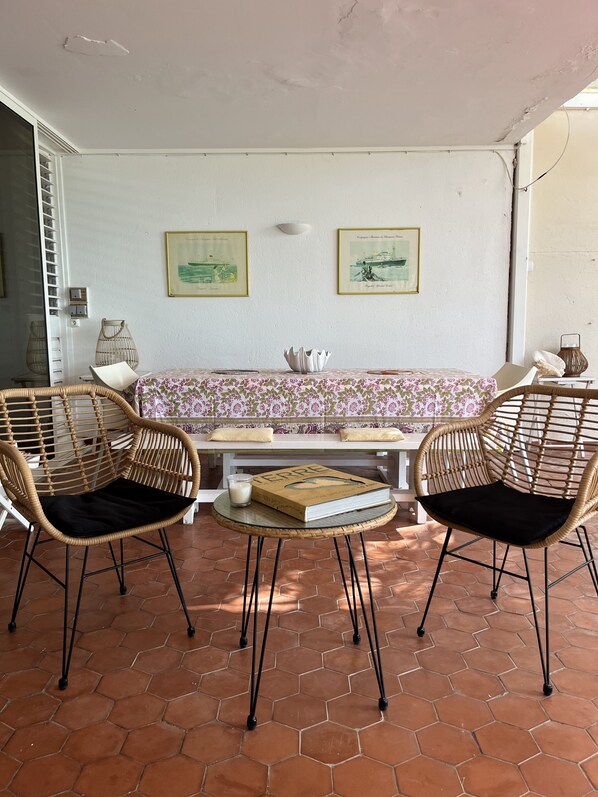 A room with a white wall, two wicker chairs, a small round table, a sofa with a floral cover, and two framed pictures.