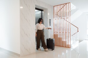 A person with a suitcase at the elevator entrance in a modern building with marble walls and a wooden staircase. - SNOW HOUSE Buriram (Buriram)
