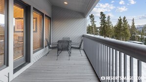 A balcony with a table and chairs, overlooking a snowy landscape with trees and mountains.