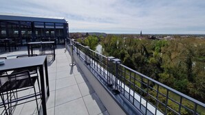 A terrace with tables and chairs, a railing, and a view of a river and distant buildings.