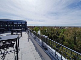 Una terraza con mesas y sillas, baranda y vista a un río y edificios a lo lejos.