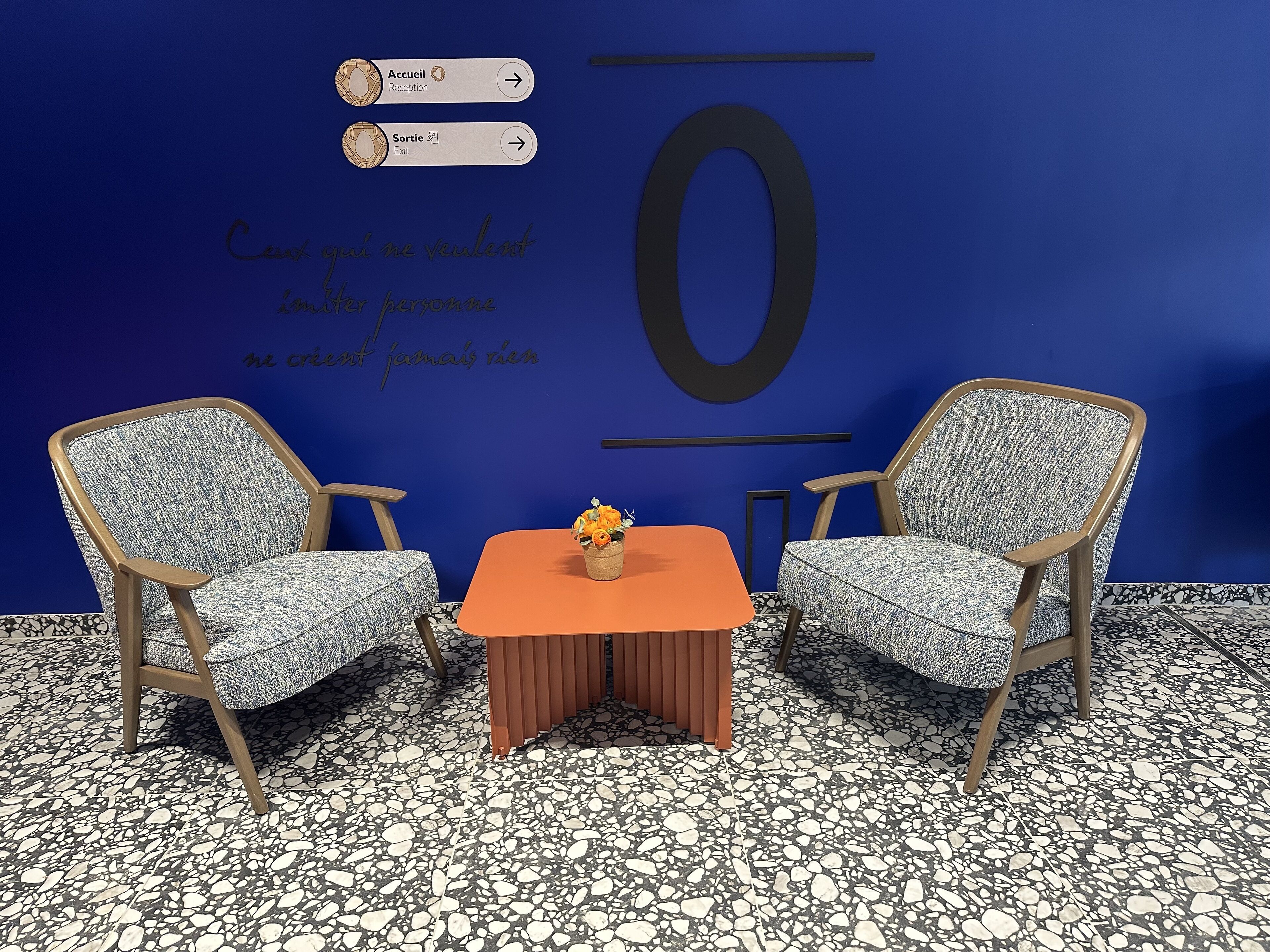 A room with two armchairs, a small table with a vase, and a wall with a large number '0' and French text.