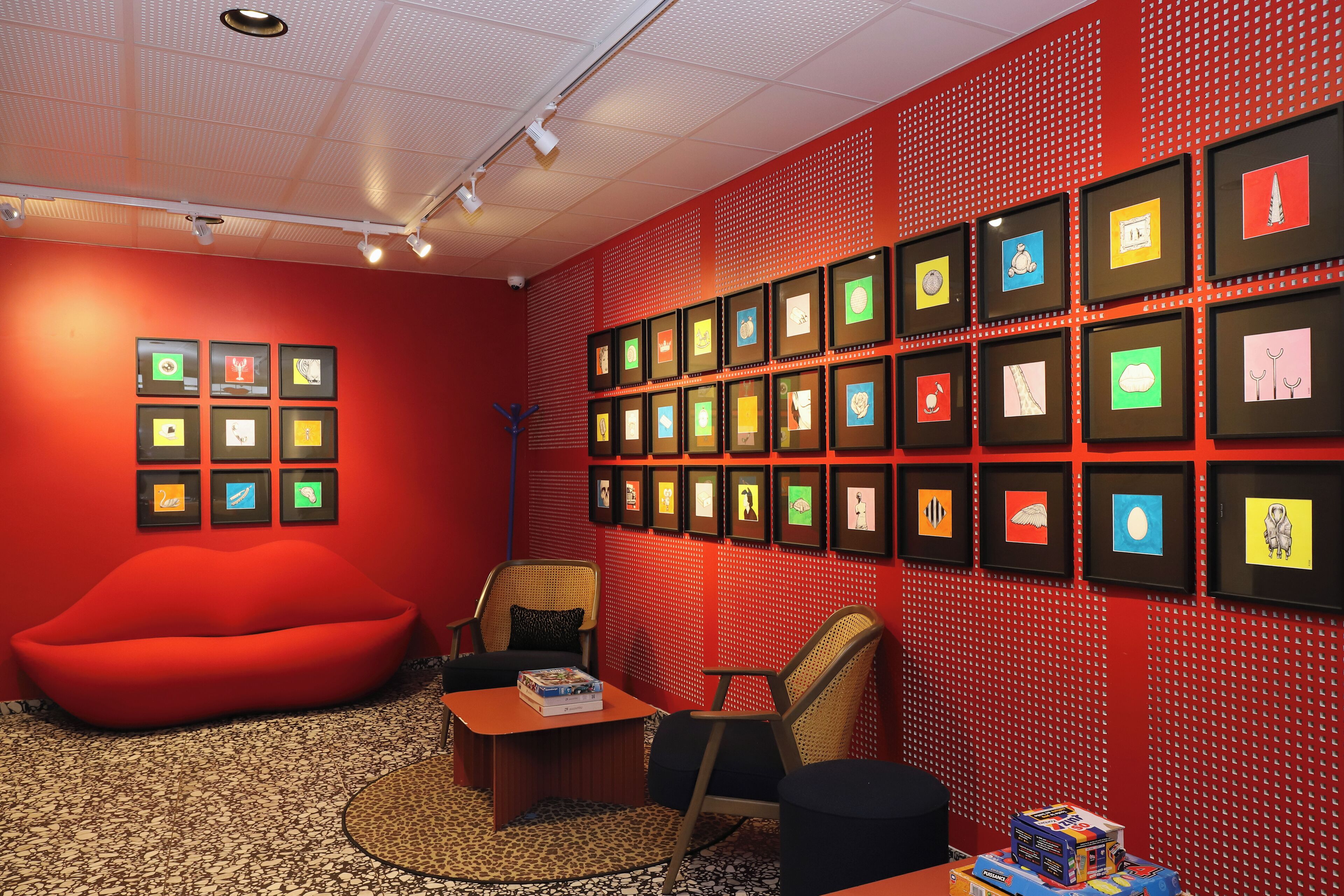 Una habitación con una pared roja, varias obras de arte enmarcadas, un sillón de rafia rojo y una pequeña mesa con libros.