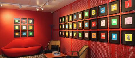 Una habitación con una pared roja, varias obras de arte enmarcadas, un sillón de rafia rojo y una pequeña mesa con libros.