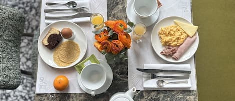 Una mesa de desayuno con platos de comida, cubiertos, una tetera y un jarrón con flores.