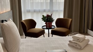 A hotel room with a bed, two armchairs, a small table with a vase, and a view of trees through sheer curtains.
