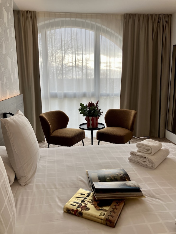 Hotelkamer met een bed, twee fauteuils, een tafeltje met een vaas en uitzicht op bomen door dunne gordijnen.