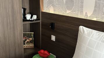 A bedroom with a bed, a nightstand, a shelf with books, a decorative wall, and a patterned wallpaper.