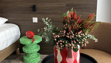 Een hotelkamer met een bed, een kruk met een groen-wit patroon, een rode en witte vaas met bloemen en een ronde zwarte tafel.
