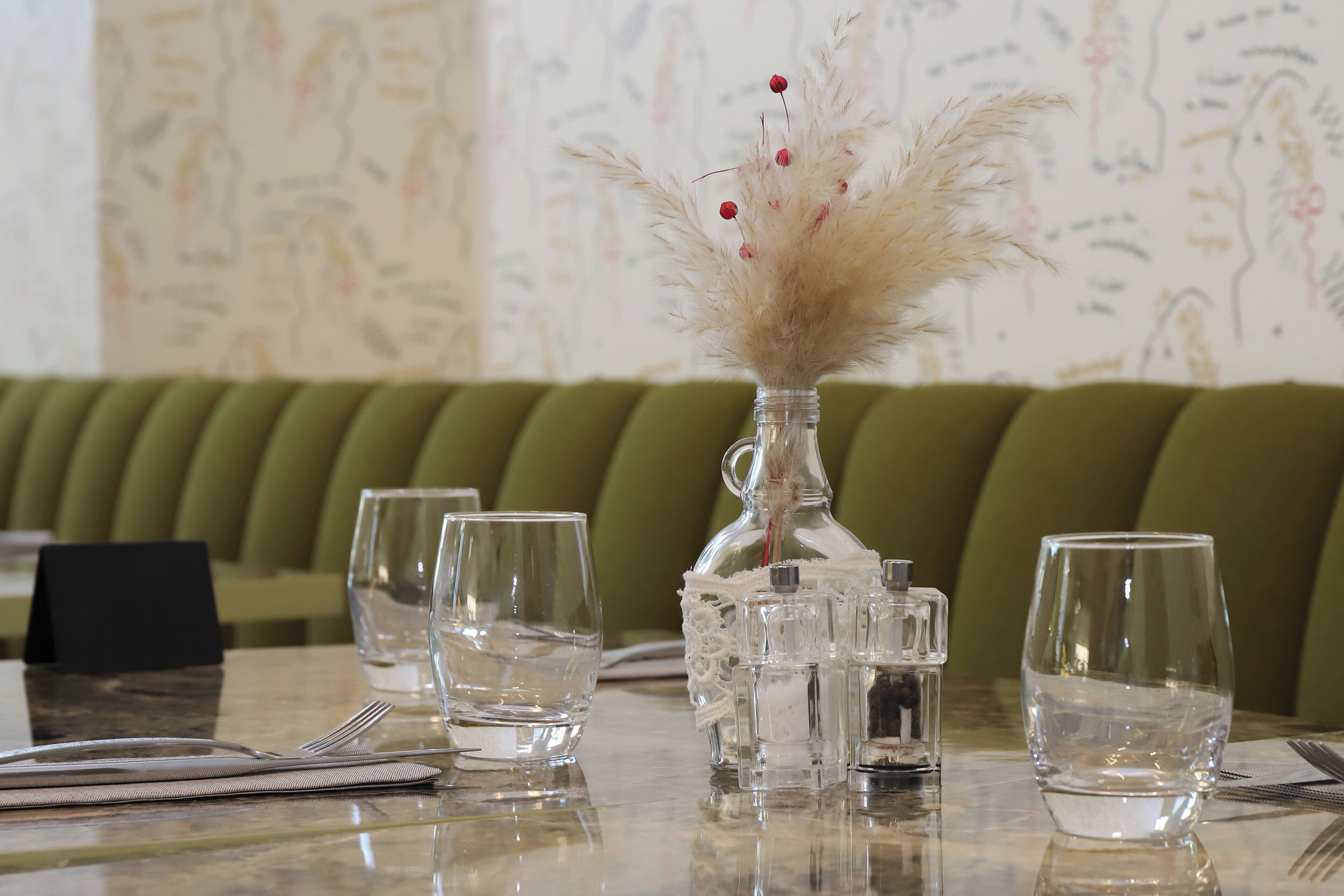 Una mesa de comedor con un jarrón de cristal vacío que contiene un arreglo de plantas secas, varios vasos vacíos y un juego de cubiertos.