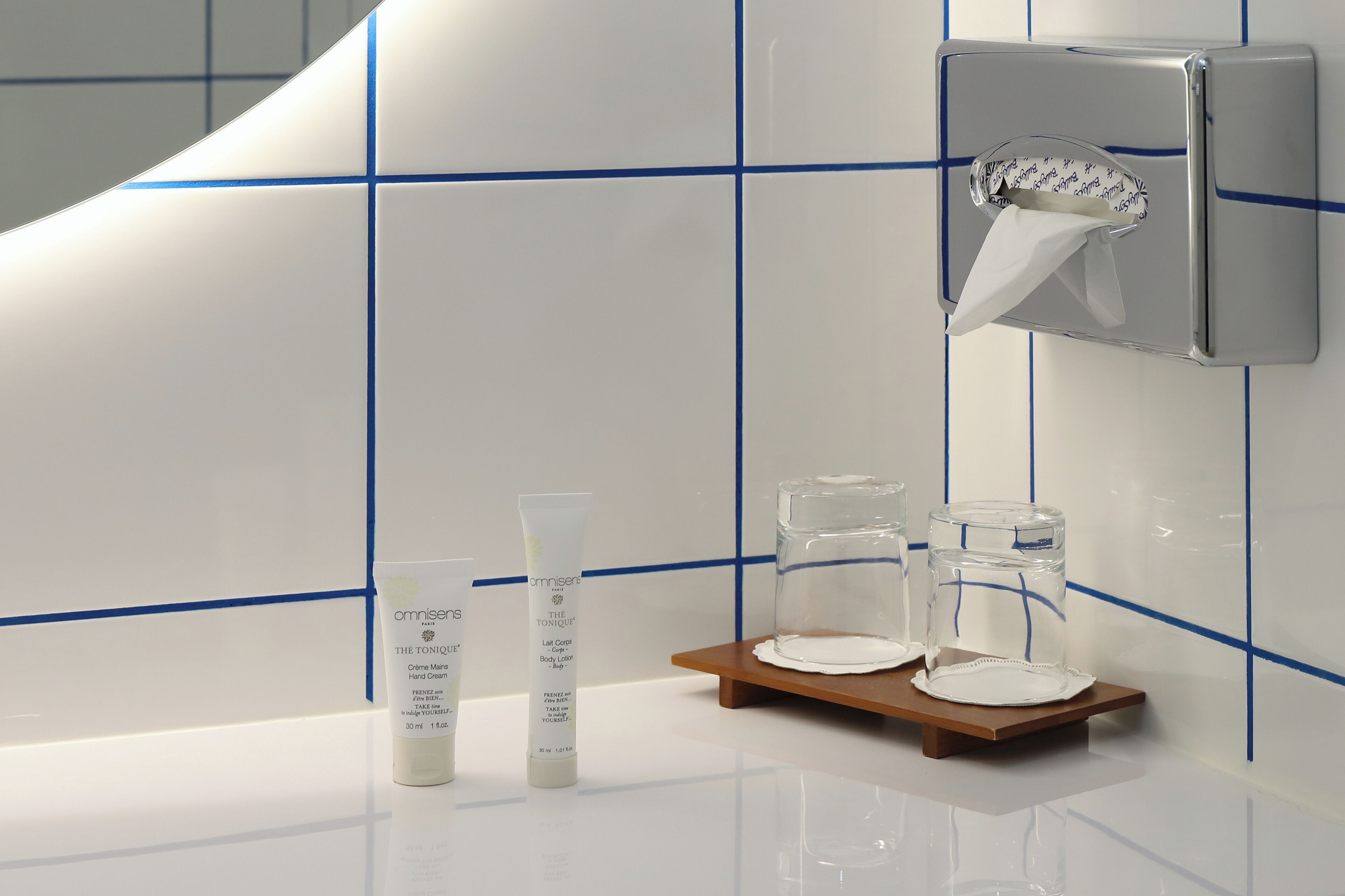 A bathroom with white tiles and blue lines, a hand soap dispenser, two glass containers, and a small wooden tray.