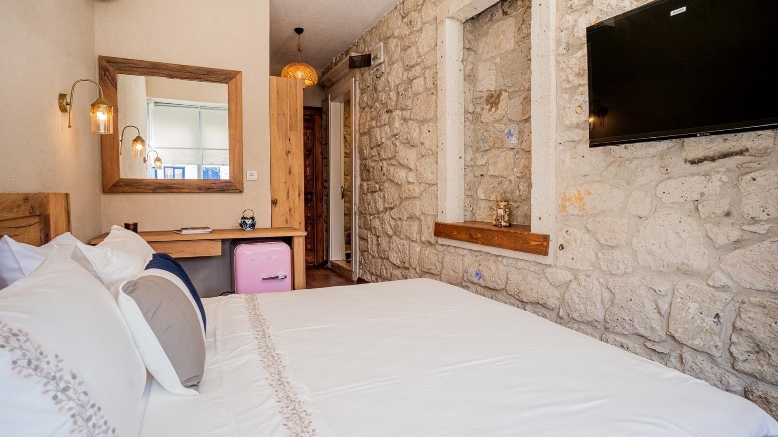 A bedroom with a large bed, a wooden dresser, a pink suitcase, a stone wall, and a TV mounted on the wall.