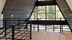 A spacious interior with a high wooden ceiling, a central circular light fixture, and large windows offering a view of trees. - One-of-a-Kind Double A Frame w/ Walkway, Windows Galore & Tons of Amenities! (Lead)