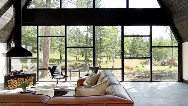 A spacious loft with a sloped ceiling, large windows offering a view of the forest, a modern chandelier, and a leather sofa. - One-of-a-Kind Double A Frame w/ Walkway, Windows Galore & Tons of Amenities! (Lead)