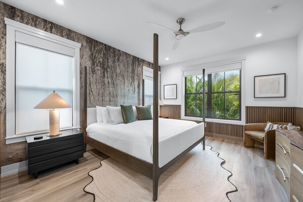 A modern bedroom with a four-poster bed, a nightstand with a lamp, a dresser, and a window with a view of greenery.