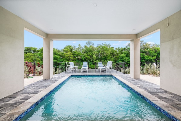 A rectangular pool with a tiled edge, surrounded by white lounge chairs and a pergola.