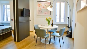 A modern kitchen with a dining area, wooden flooring, a flat-screen TV, and a large window providing natural light.