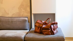 A brown leather handbag on a grey cushioned bench in a room with a textured wall and a plain white wall in the background.