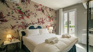 A hotel room with a floral wallpaper, a bed with white linens, a nightstand with a lamp, and a view of the outdoors through a sliding door. - Zagara House - Villa TerraFORTE (Marina di Ragusa)