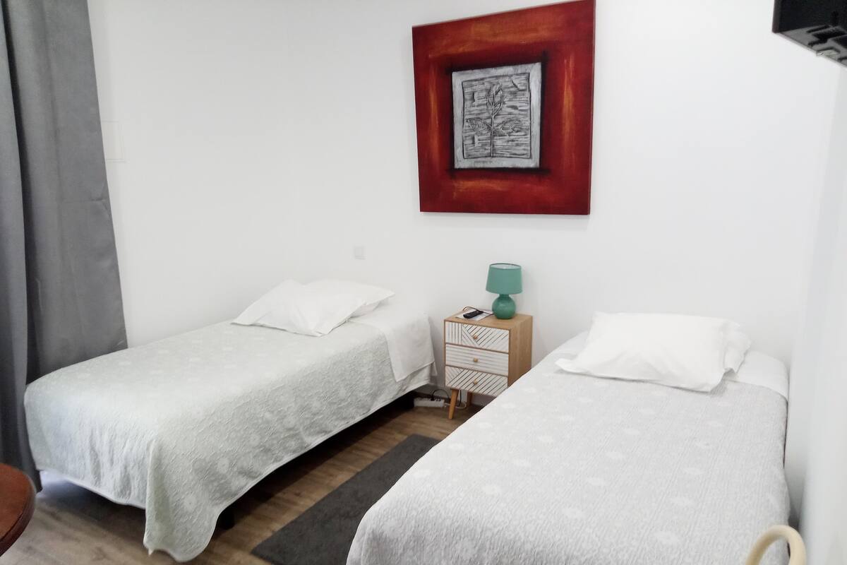Una habitación con dos camas individuales, una mesita de noche con lámpara y un cuadro enmarcado en la pared.