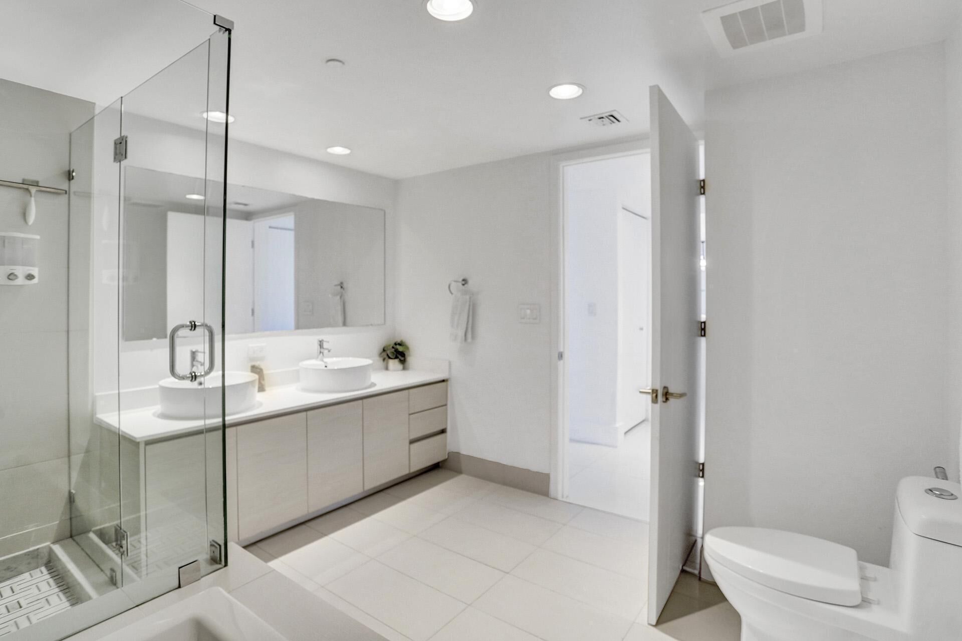 Un baño moderno con cabina de ducha de vidrio, dos lavamanos, inodoro y un espejo grande.
