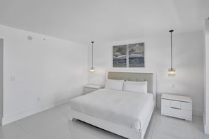 A minimalist bedroom with a large bed, two bedside tables, and a ceiling-mounted artwork. - Luxury Condo at Beach Walk Resort (Hallandale Beach)