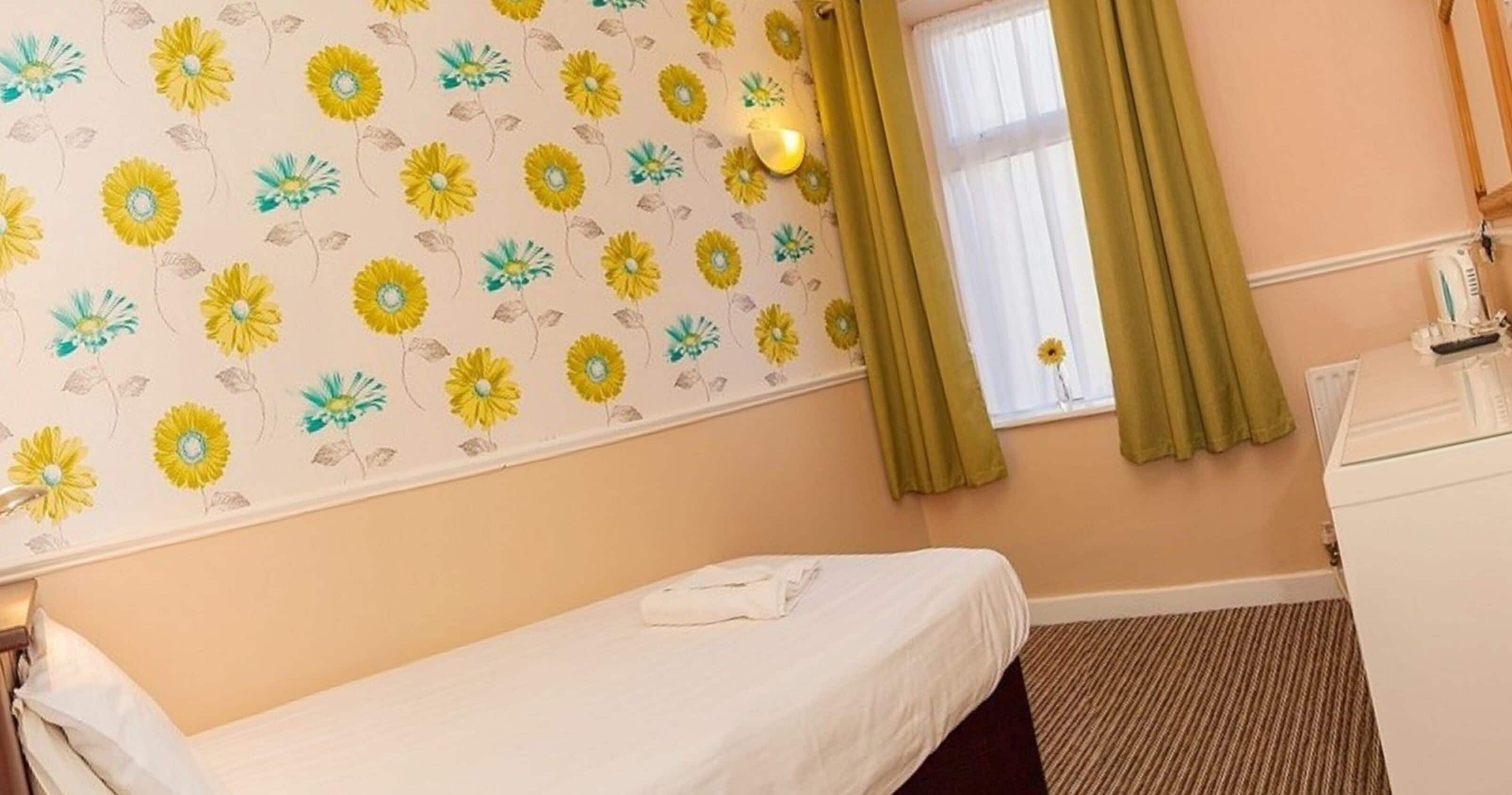 Ein Schlafzimmer mit einem Einzelbett, einem Fenster mit Vorhängen, einem Heizkörper und einem kleinen Tisch mit Fernbedienung.