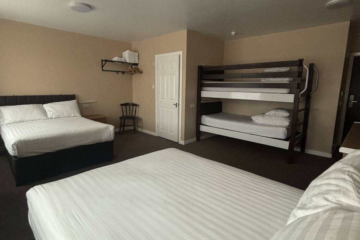 Une chambre d’hôtel avec un grand lit, un lit plus petit, un lit superposé, une chaise, une étagère avec des serviettes et une porte.