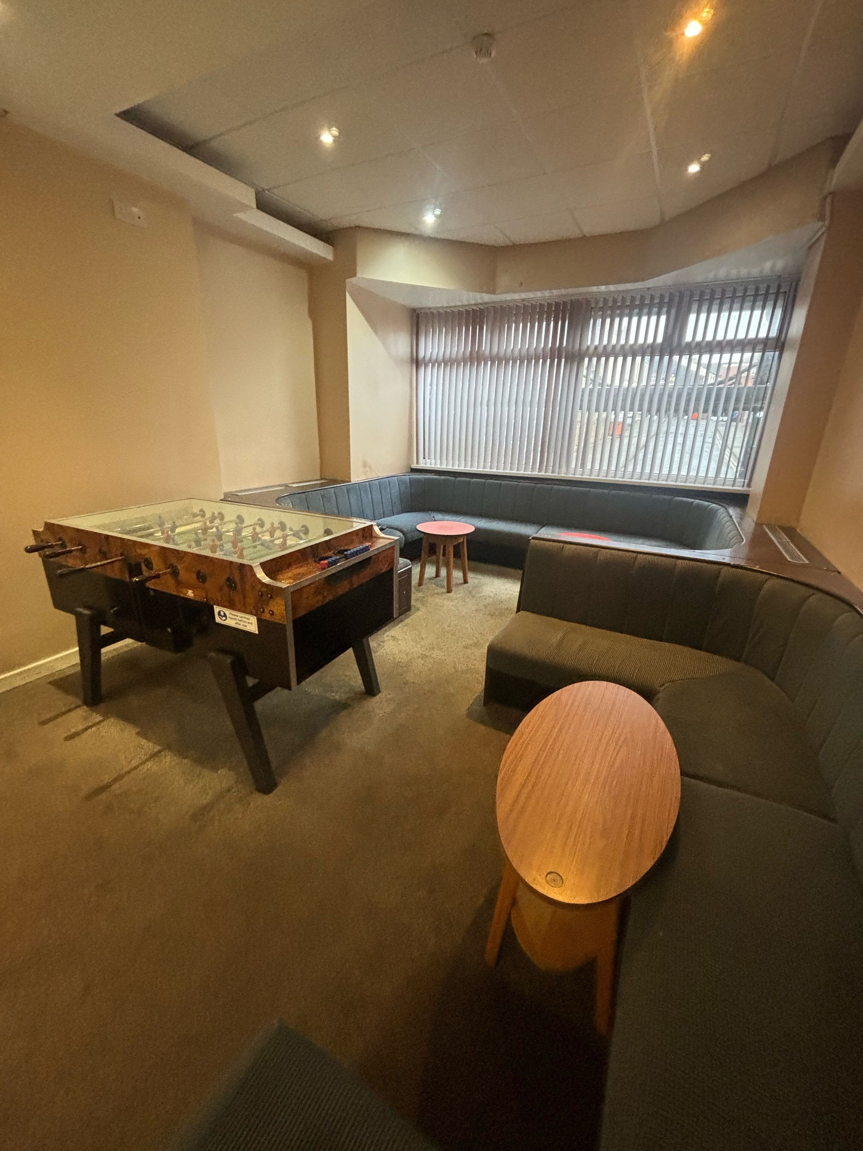 A room with a foosball table, a sofa, a small round table, and a window with blinds.
