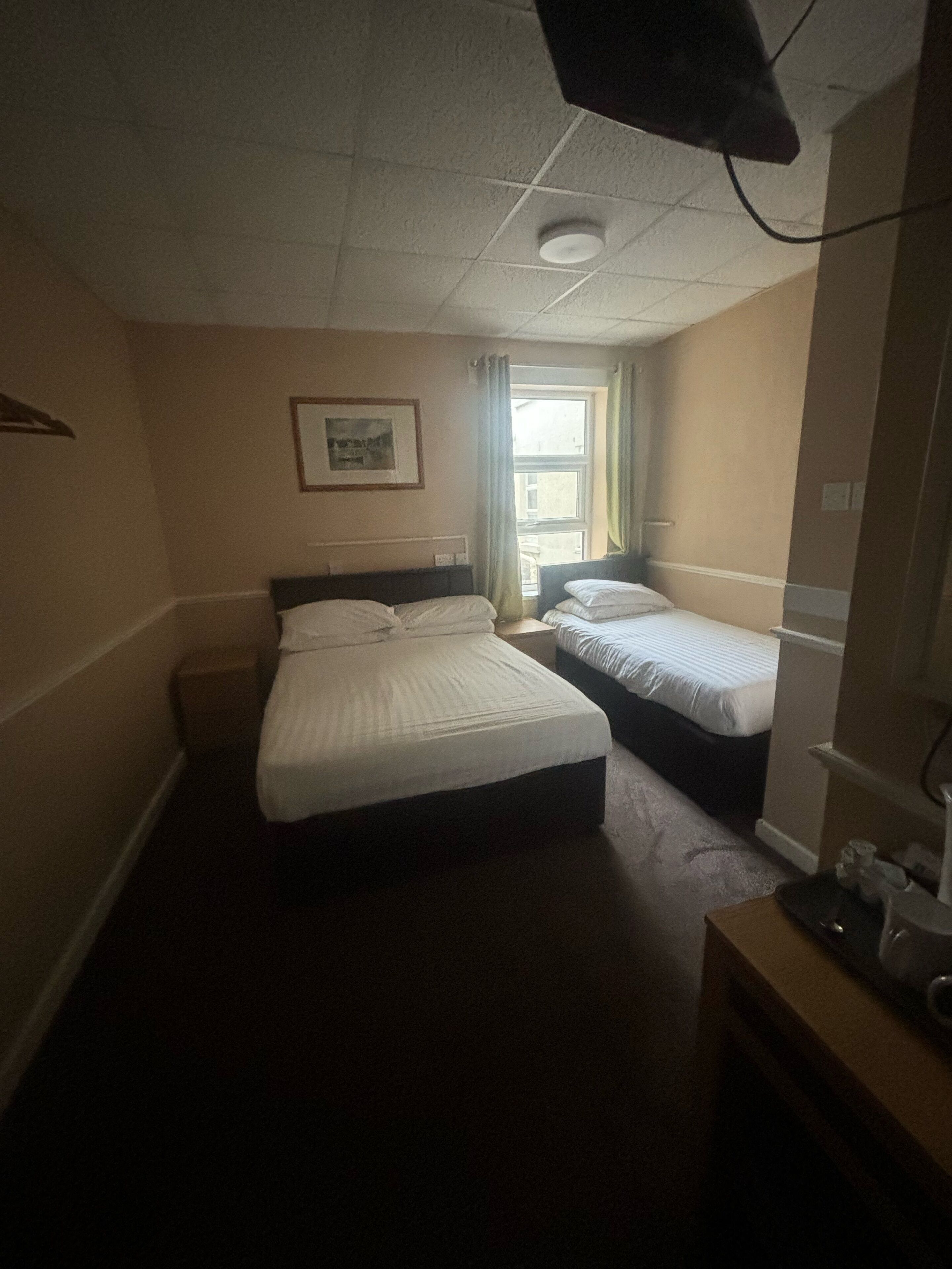 A hotel room with two single beds, a small desk, a window with curtains, and a ceiling light.