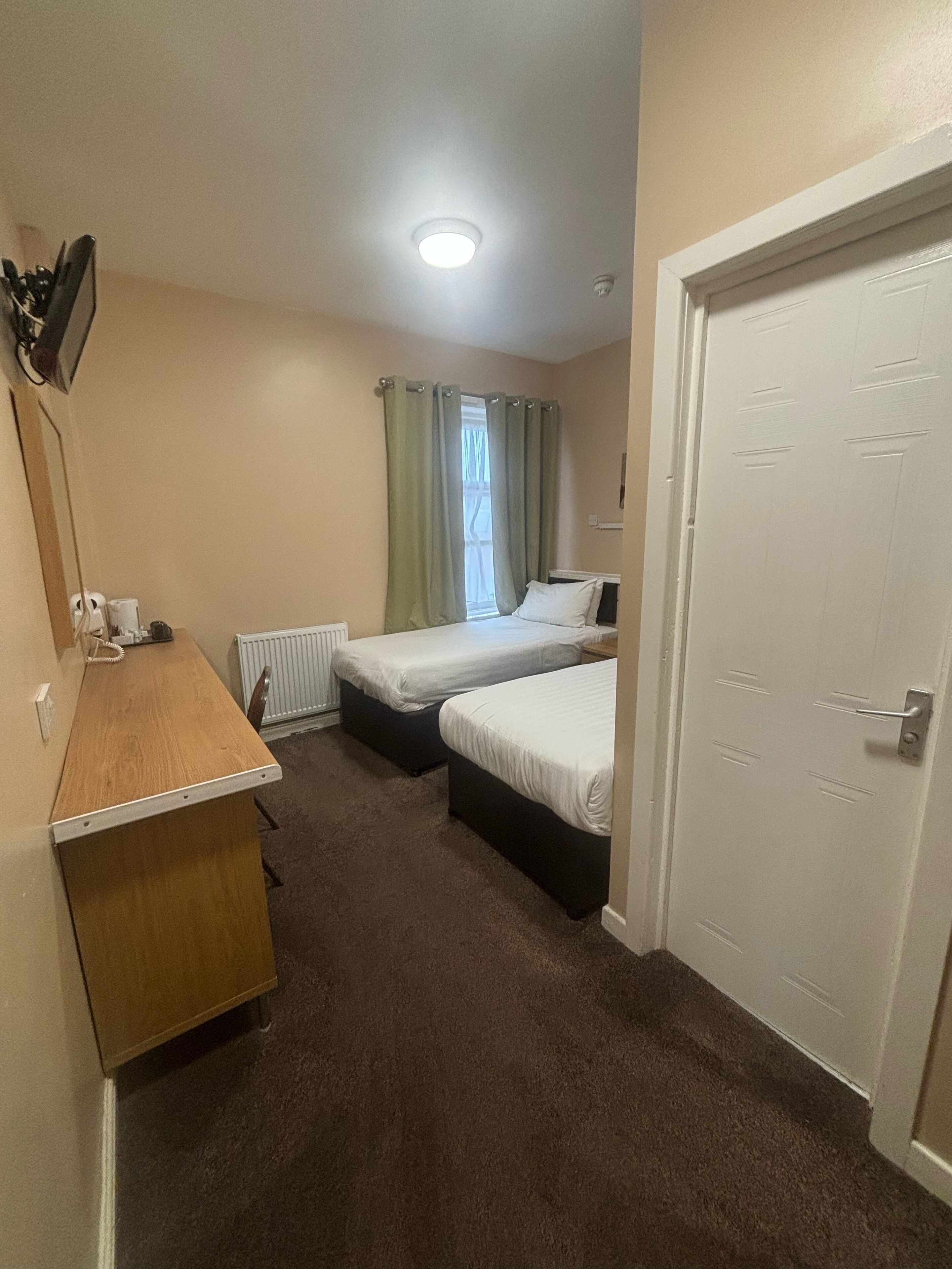 Een kleine, compacte kamer met twee eenpersoonsbedden, een houten bureau, een radiator en een deur.