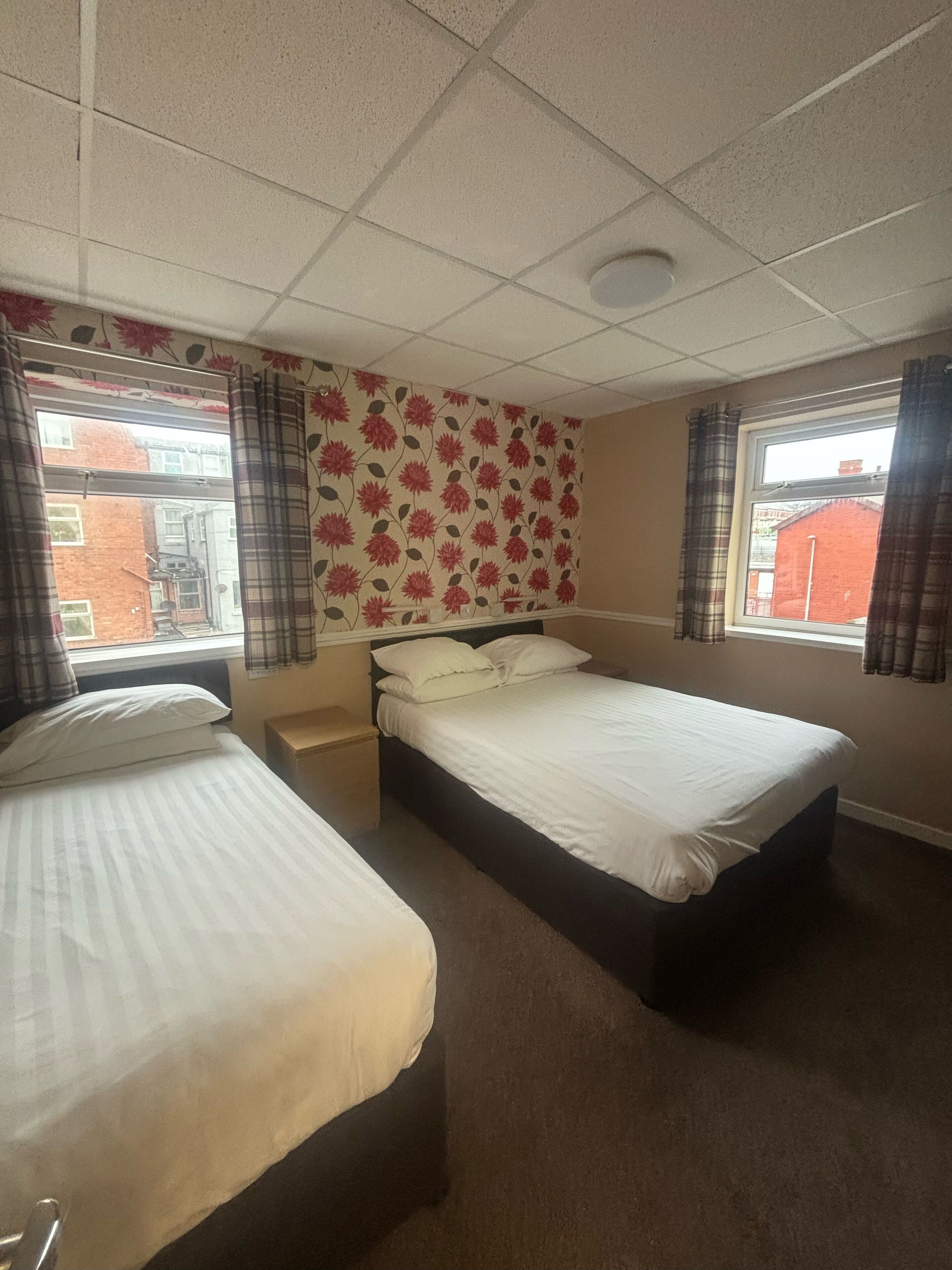 A hotel room with two single beds, a headboard, a small bedside table, a window with curtains, and a floral-patterned wall.