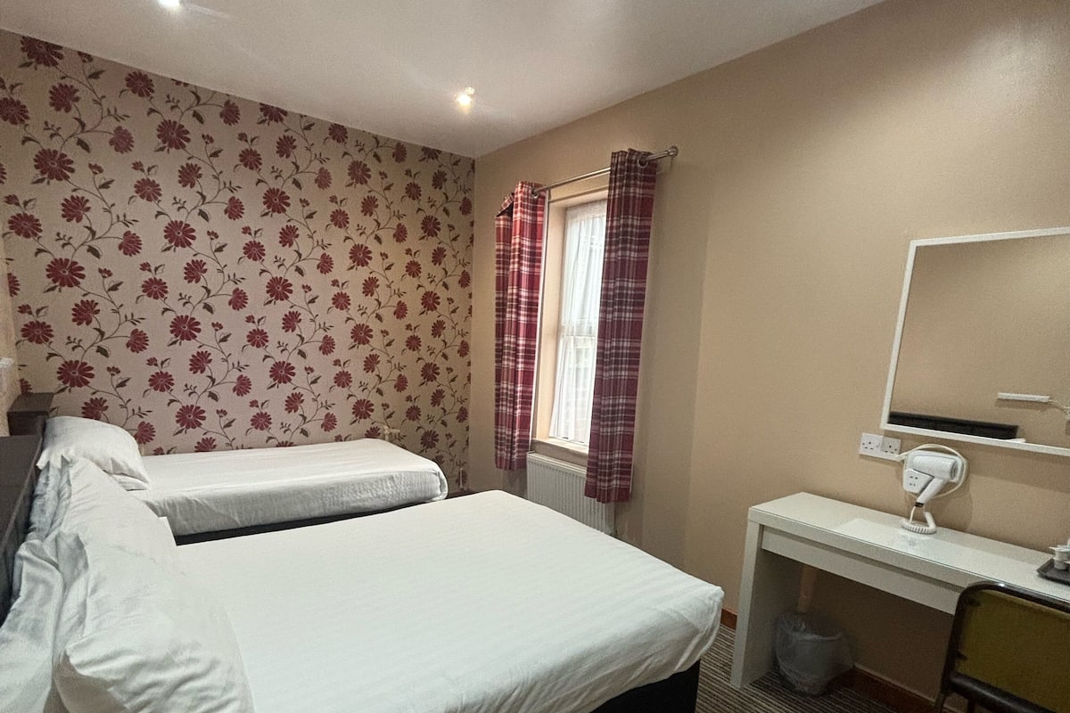Une chambre d’hôtel avec deux lits, un bureau avec une chaise, un radiateur et une fenêtre avec des rideaux.