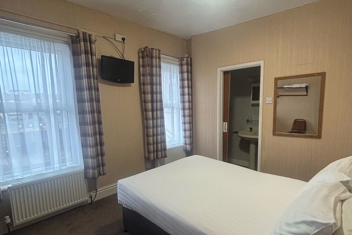 Une chambre d’hôtel avec un lit simple, un radiateur, un téléviseur fixé au mur et une porte donnant sur une salle de bain.