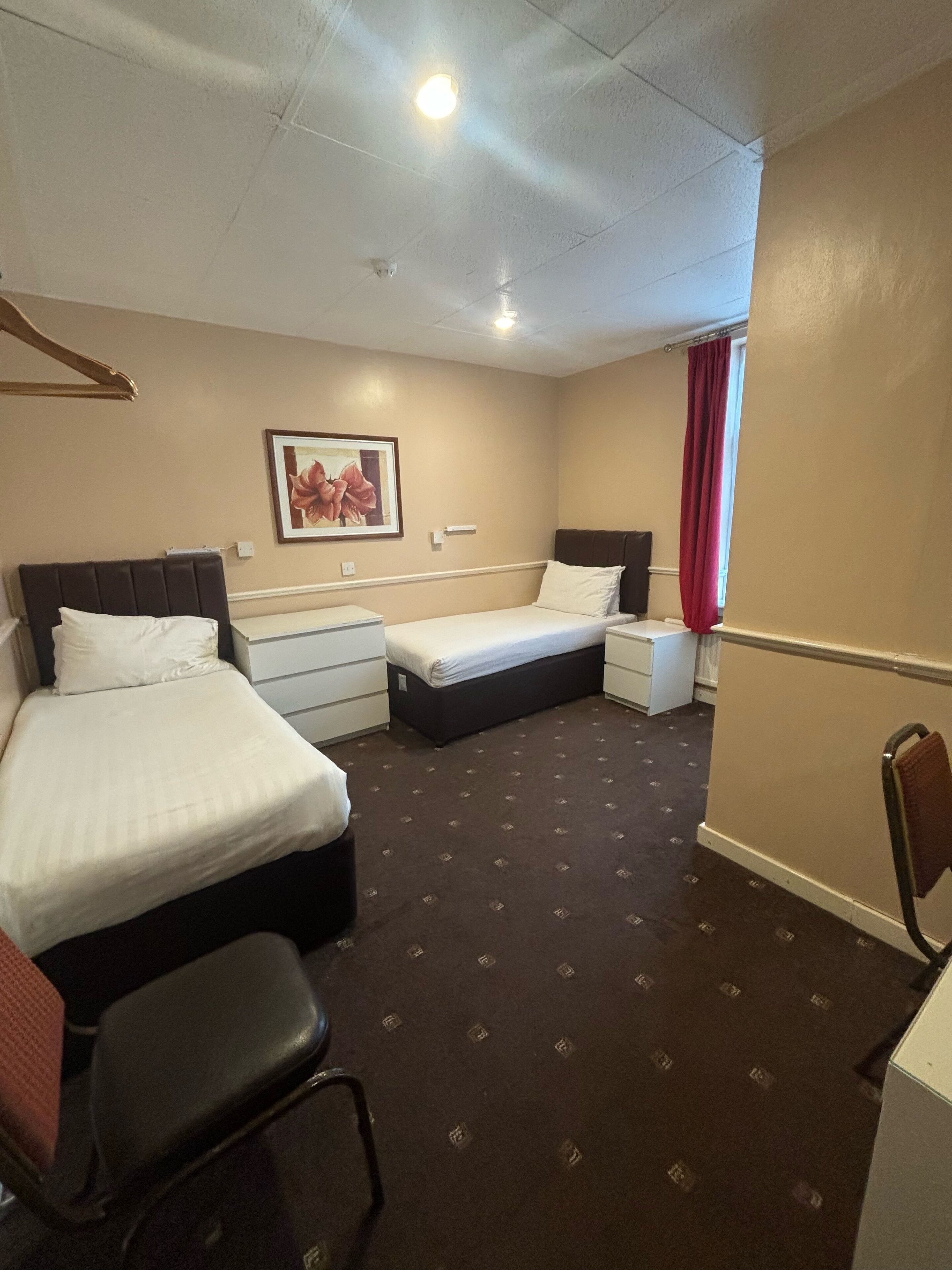 Une chambre d’hôtel avec deux lits, un bureau, une chaise, un rideau rouge et un tableau encadré au mur.