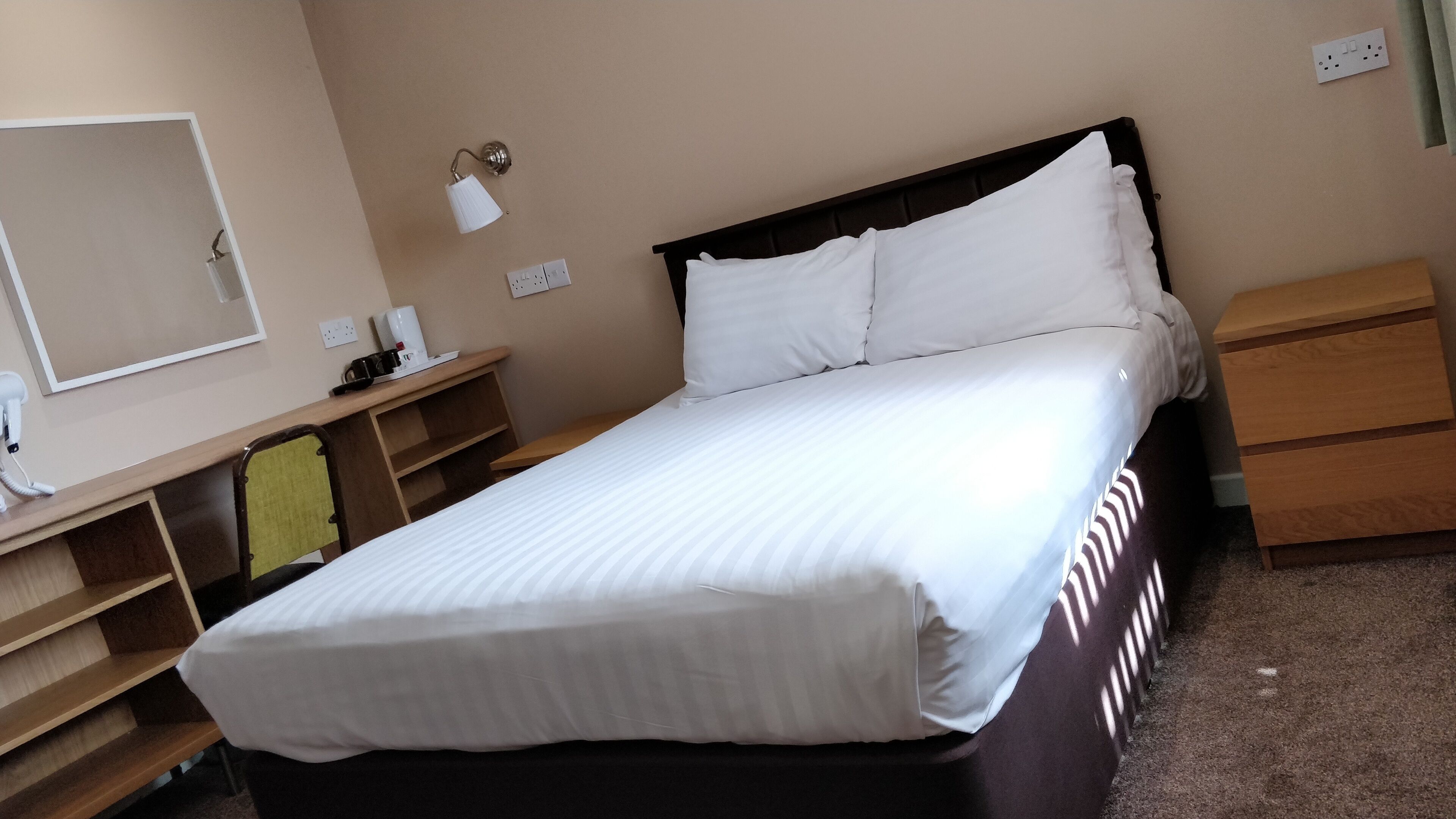 Una cama bien hecha con ropa blanca, una mesita de noche con lámpara, una pequeña mesa con cafetera y un aparador de madera.