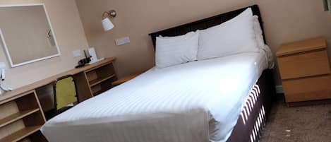 Una cama bien hecha con ropa blanca, una mesita de noche con lámpara, una pequeña mesa con cafetera y un aparador de madera.