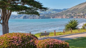 Un parco costiero con una panchina, cespugli fioriti e vista sull'oceano e sulle colline.